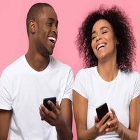 man and woman black young couple using cell phones sitting side by side laughing. pink background