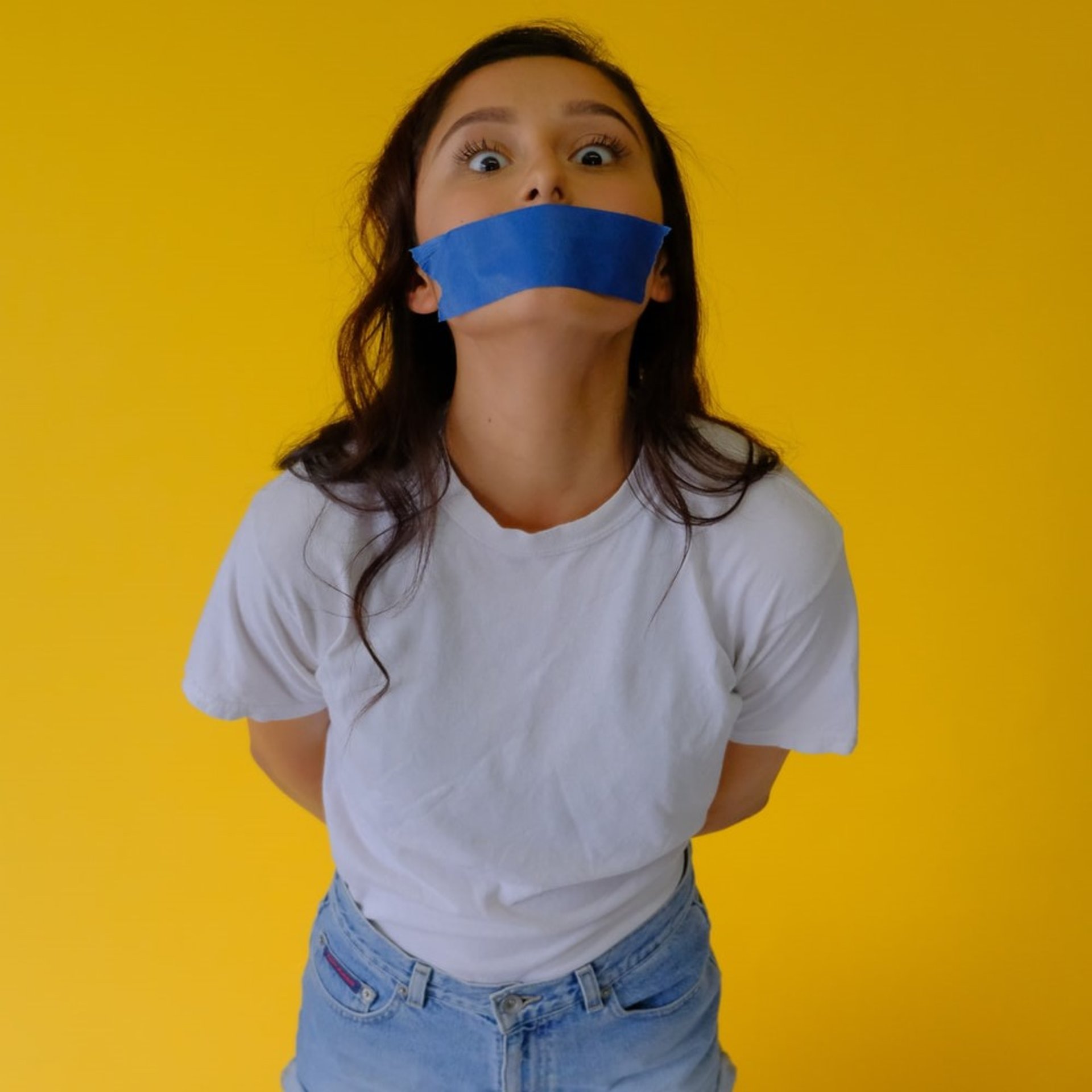 woman restrained with hands behind back, with blue tape on mouth, in front of yellow background